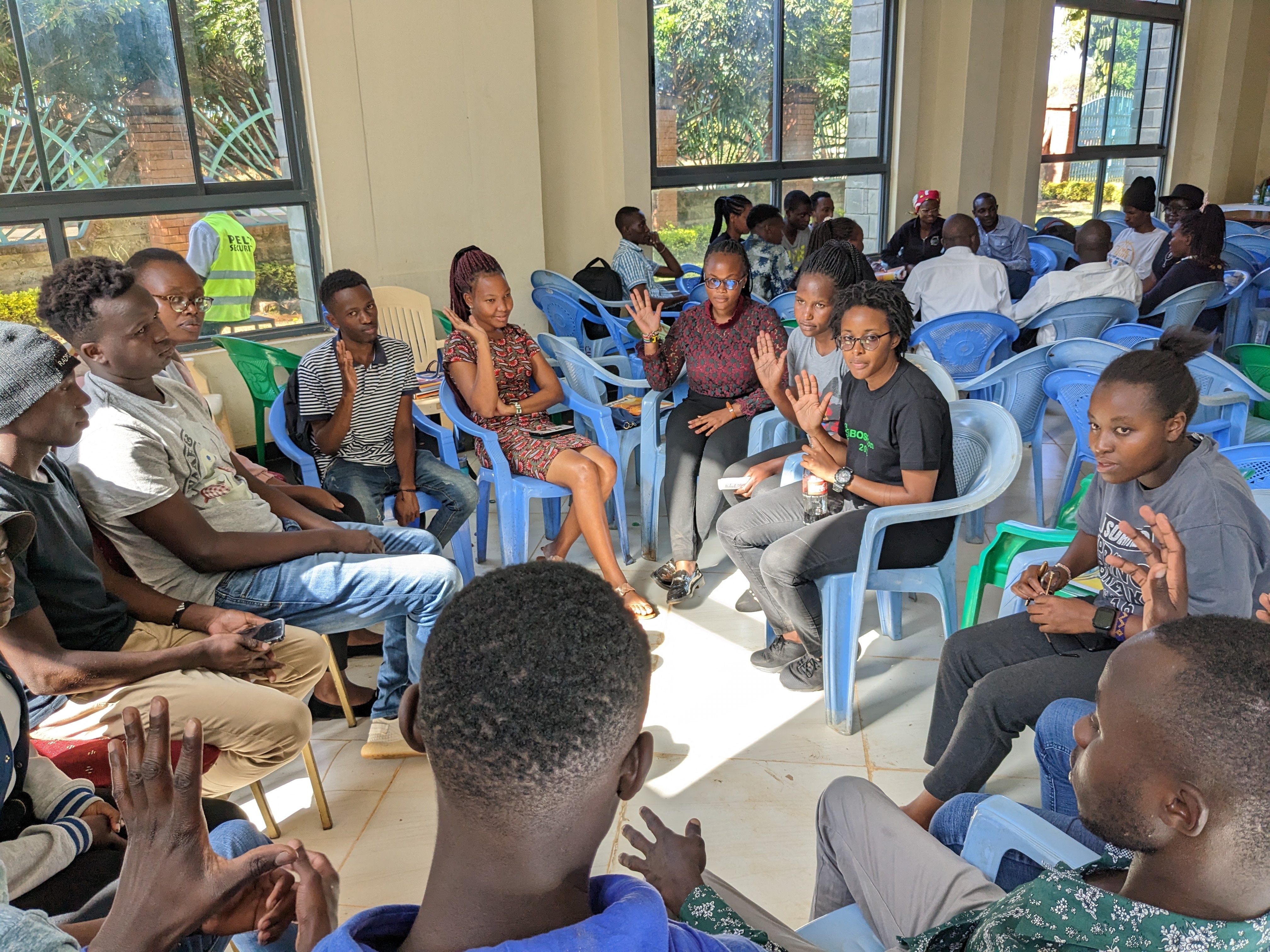 Bioinformatics Hub of Kenya members, young students, approximately 6 women and five men, sitting in blue chairs forming a circle, inside a modern concrete building with high ceilings, many of them raising their hands as in voting, engaging in a conversation, the sunlight creates a square shape in the middle of the circle (BHKi)