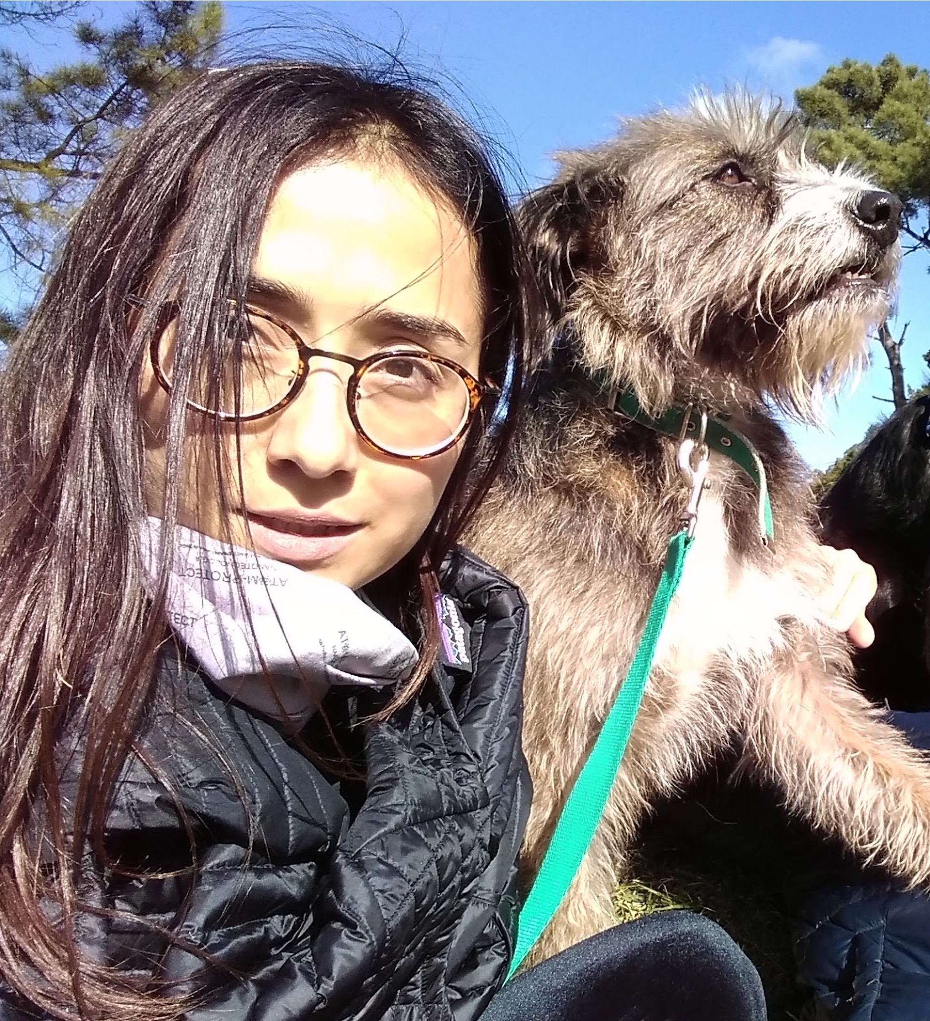 Paz is a woman from Chile. She has long black hair. She is wearing a black winter jacket and a pair of rimmed glasses, and holding her pet, a furry dog who seems to be enjoying sunlight