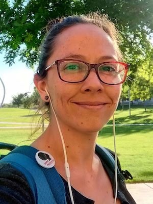 Andrea is a woman from Argentina. She has tied back her hair. She is wearing a pair of red rimmed glasses, has her white earbud - probably listening to music on a sunny day. She is wearing a blue T shirt and a backpack. She is smiling and seem to be enjoying a pause from her walk to take a selfie.