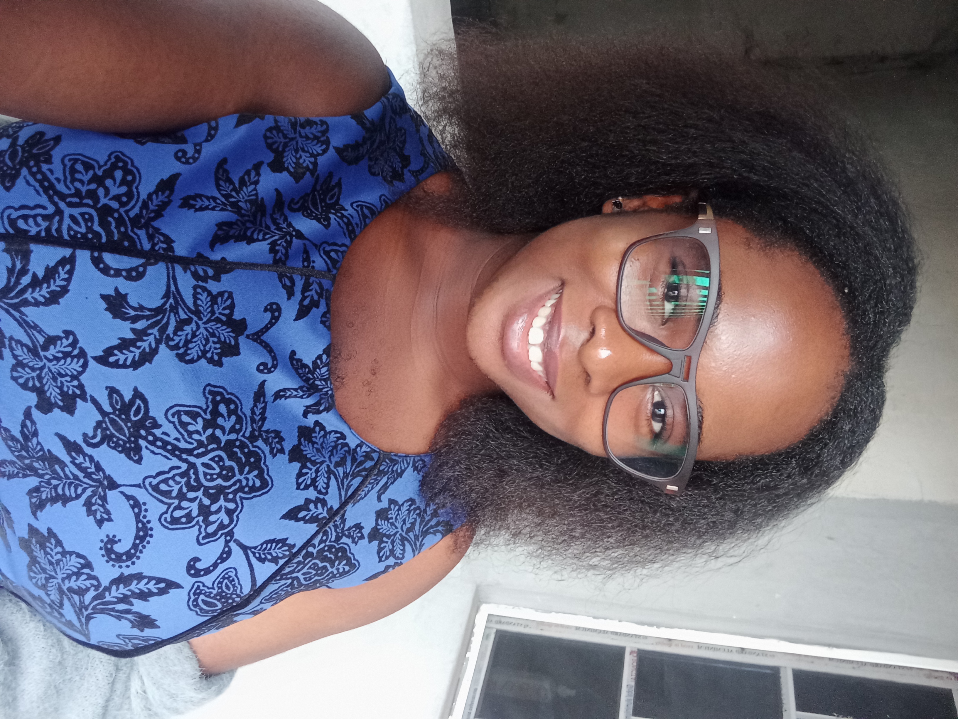 A photo of Debs, a woman from Nigeria. She has rich, black hair and is wearing a blue sleeveless dress, a pair of glasses and a smile.